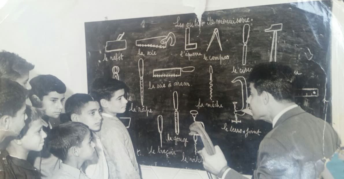 Archival photograph of a carpentry class, instructor teaching hand tools at a blackboard